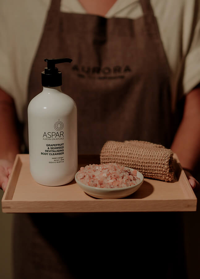 Person holding a tray with a bottle of Aspar skincare product, a bowl of sea salt, and a loofah.