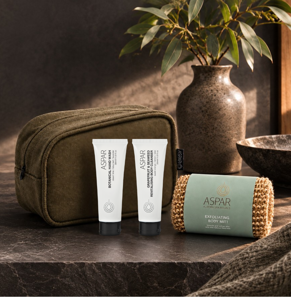 A travel set from Aspire and Grooming featuring an olive green zippered pouch, two white squeeze tubes, and a rolled-up mint green and gold tube, arranged on a dark stone surface next to a ceramic vase with green leaves.