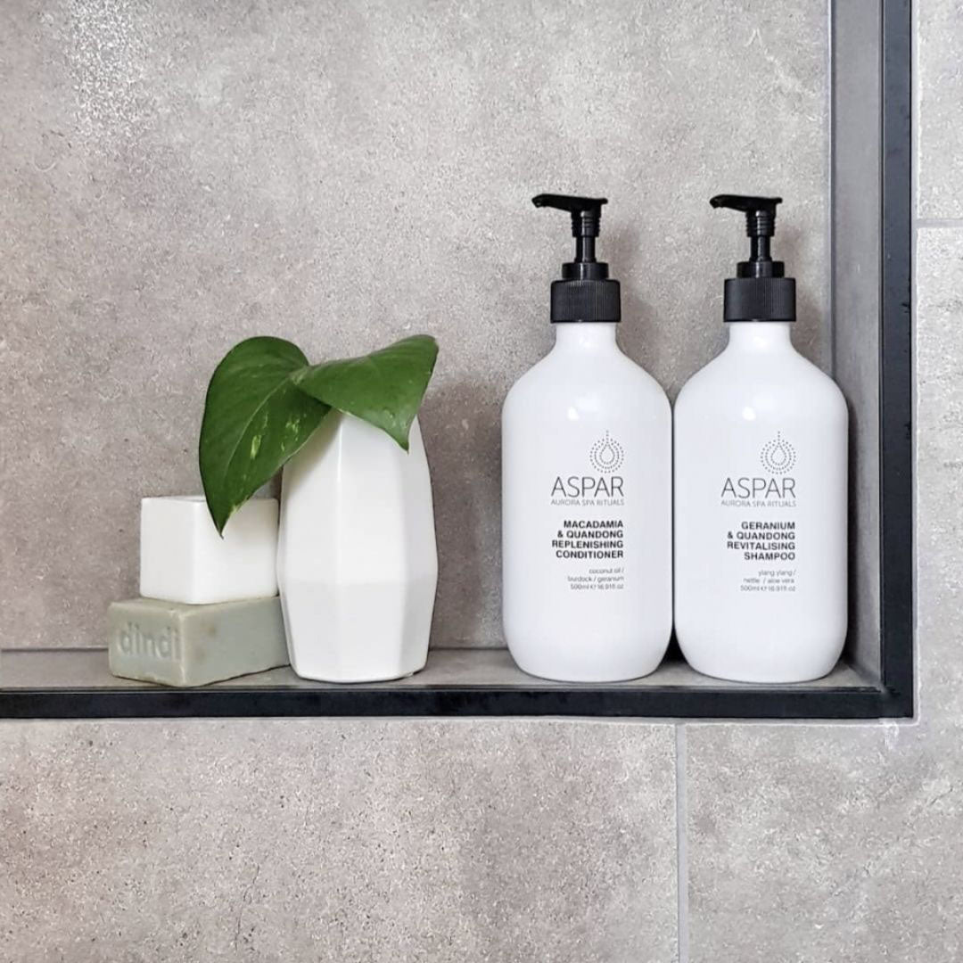 Two white bottles with black pumps labeled 'ASPAR' on a gray shelf with a plant and soap.