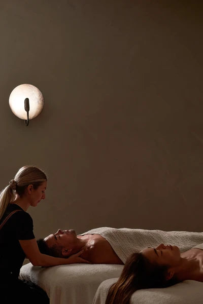 Two people receiving a massage in a dimly lit room with a wall lamp.