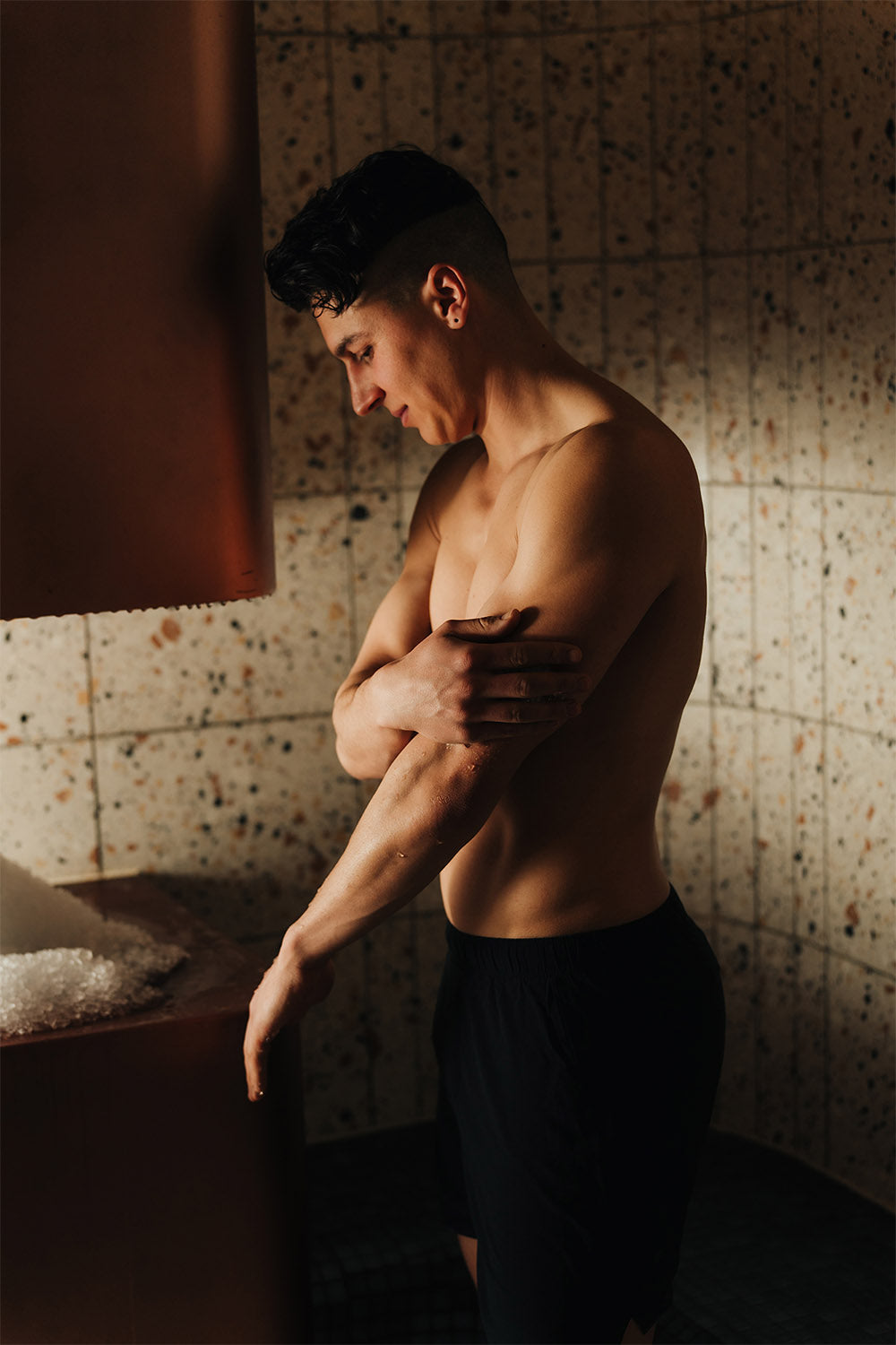 Man with a muscular build standing in a dimly lit room with textured walls.