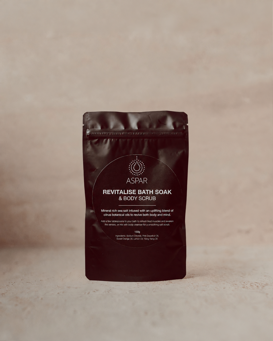 A stand-up pouch of ASPAR Revitalise Bath Soak & Body Scrub in a dark brown color, set against a neutral beige textured background.
