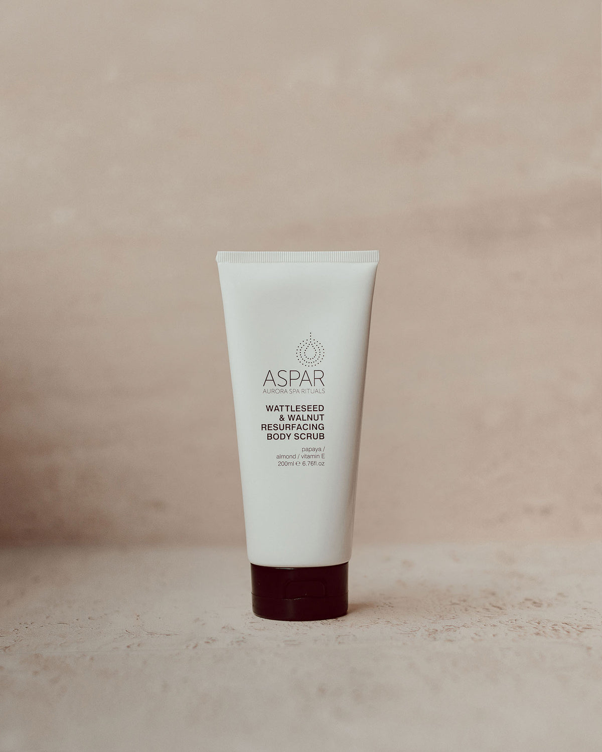 A white squeeze tube of ASPAR Wattleseed  & Walnut Resurfacing Body Scrub, standing upright on a textured beige surface against a matching beige background