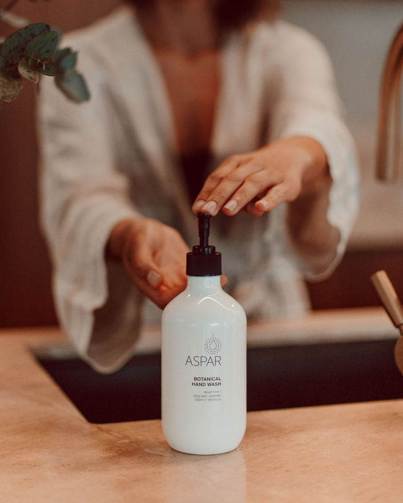 A woman in a white robe smiles while pressing the pump of an ASPAR Botanical Hand Wash on a wooden countertop in a warm, modern kitchen setting.