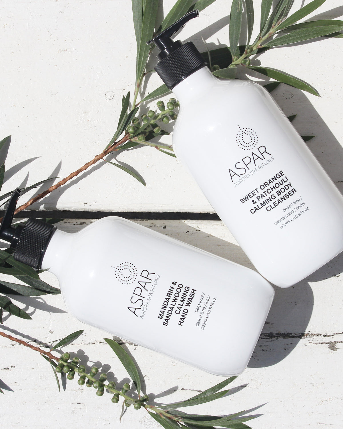 Two white ASPAR skincare bottles, one containing Deep Cleansing Clay Mask and the other Deep Pore Treatment Mask, arranged diagonally on a white wooden surface with green olive branches.