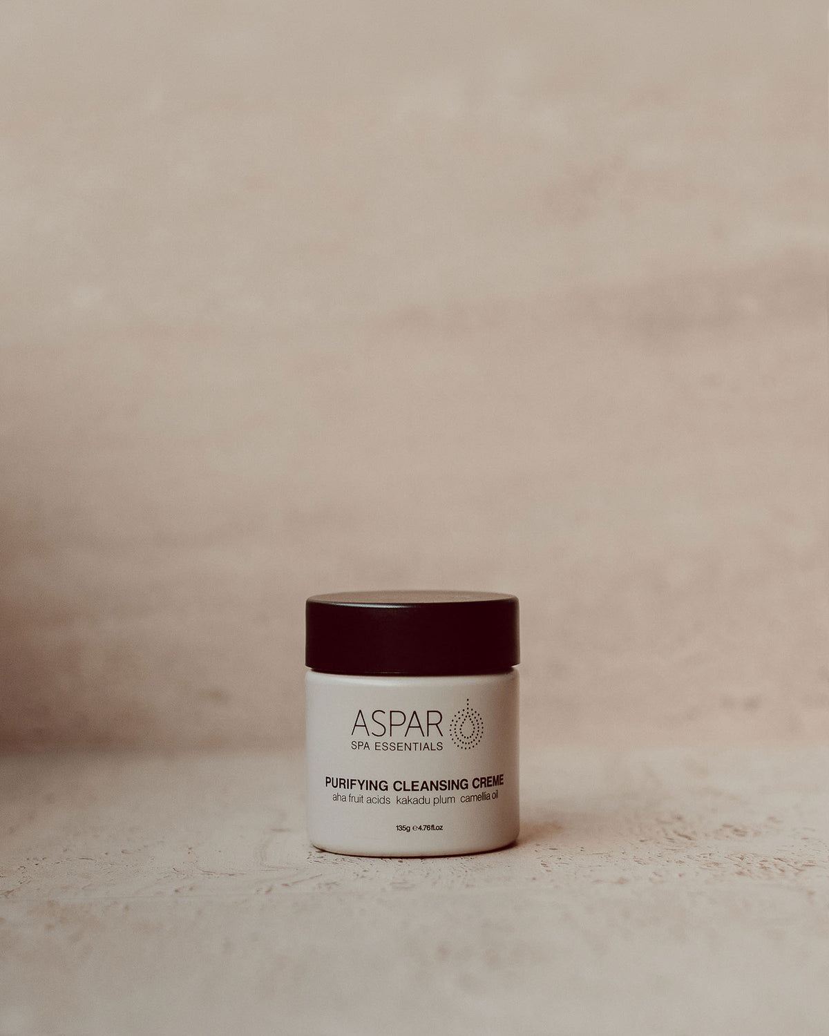 A small white jar of ASPAR Spa Essentials Purifying Cleansing Cream with a dark burgundy lid, set against a neutral beige textured background.