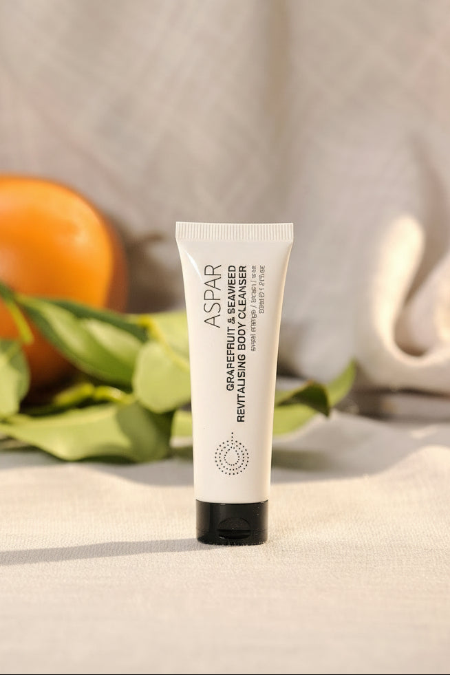 A white pump bottle of ASPAR Grapefruit & Seaweed Revitalising Body Cleanser stands next to a matching white tube with a black cap, styled with birch logs, green leaves, and an orange on a light beige fabric background.