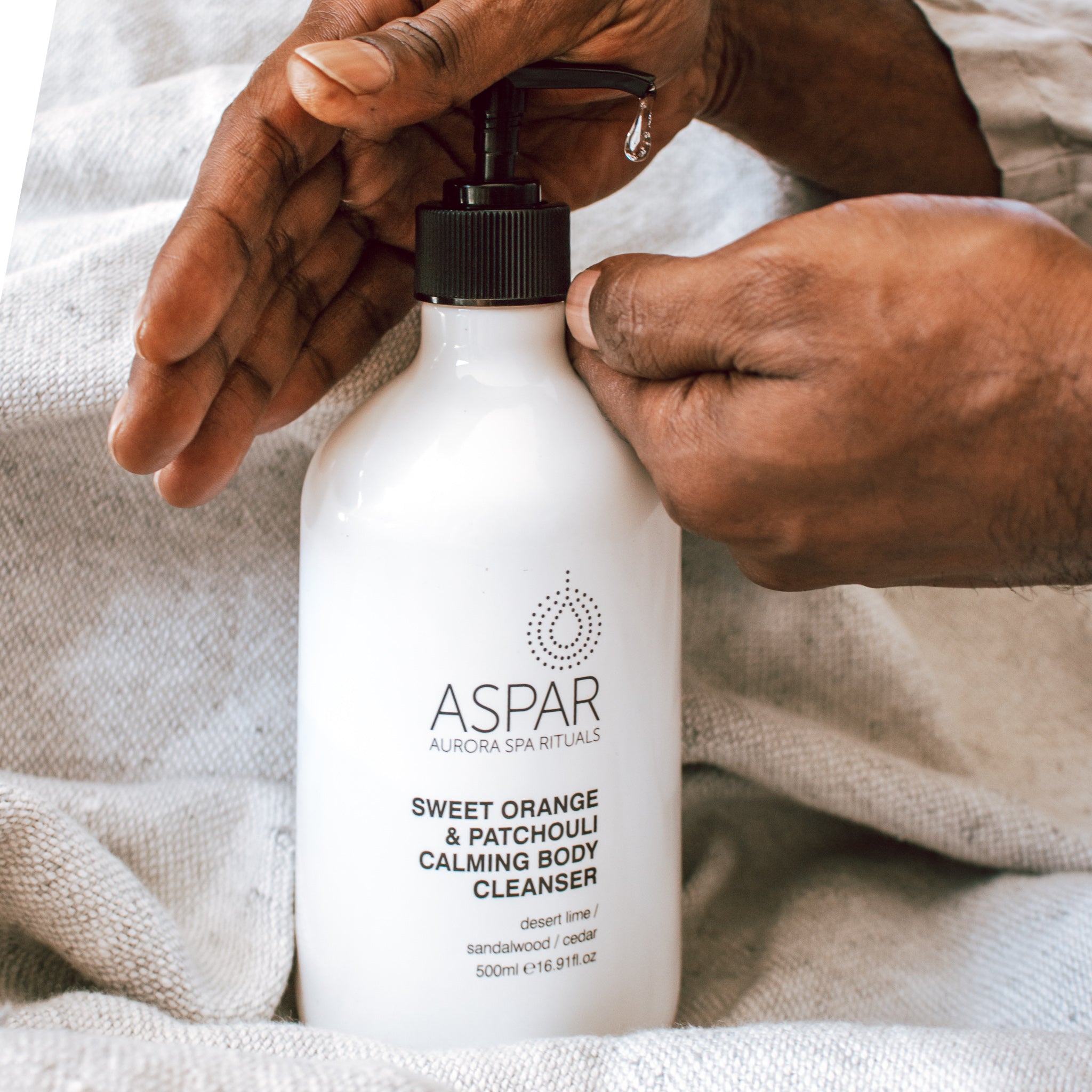 Person holding a bottle of Aspar Sweet Orange & Patchouli Calming Body Cleanser with a beige towel in the background.