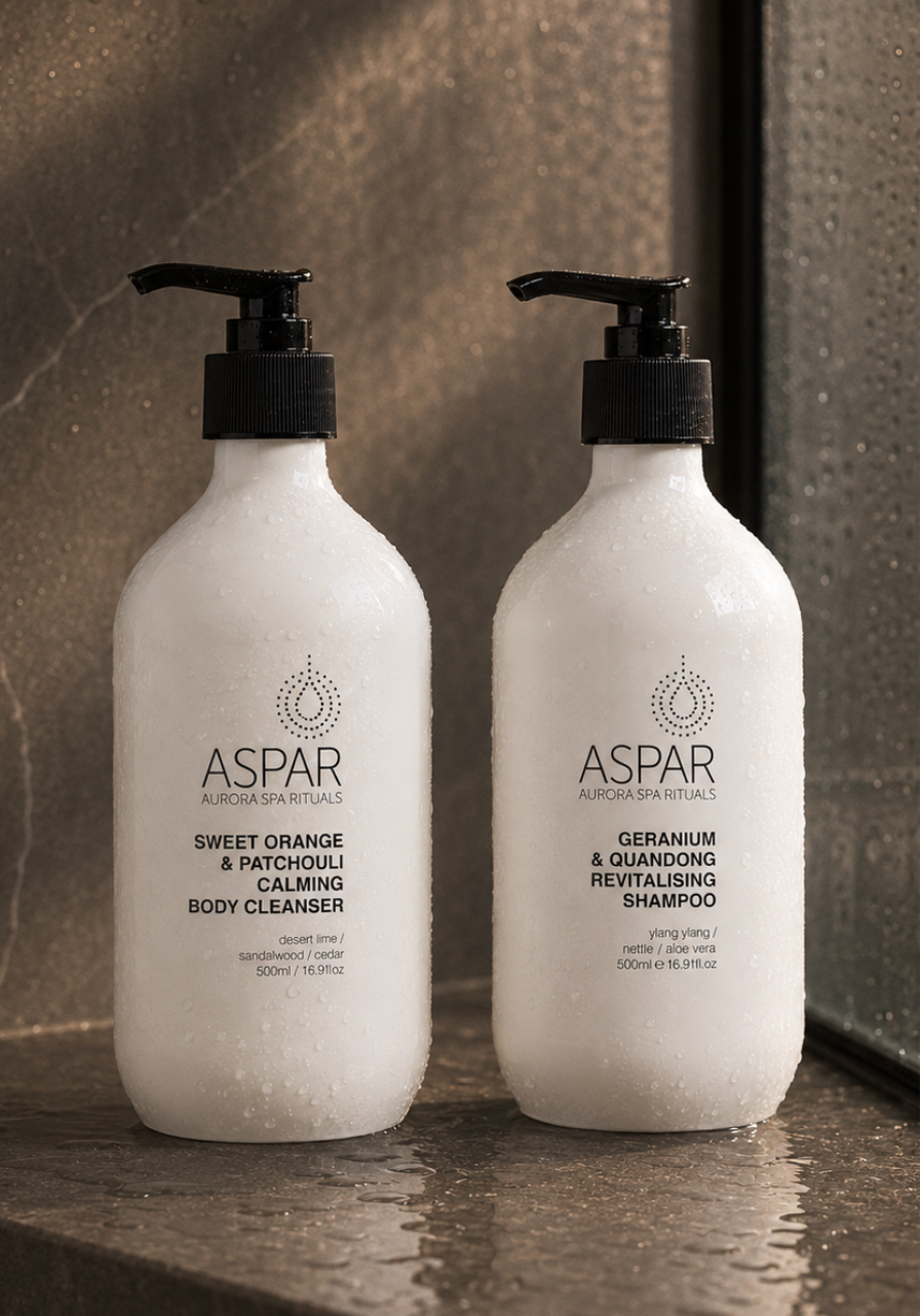 Two ASPAR luxury body care bottles on a wet stone surface in a bathroom setting. The left bottle is labeled 'Sweet Orange & Patchouli Calming Body Cleanser' and the right bottle is labeled 'Geranium & Quandong Revitalising Shampoo'.