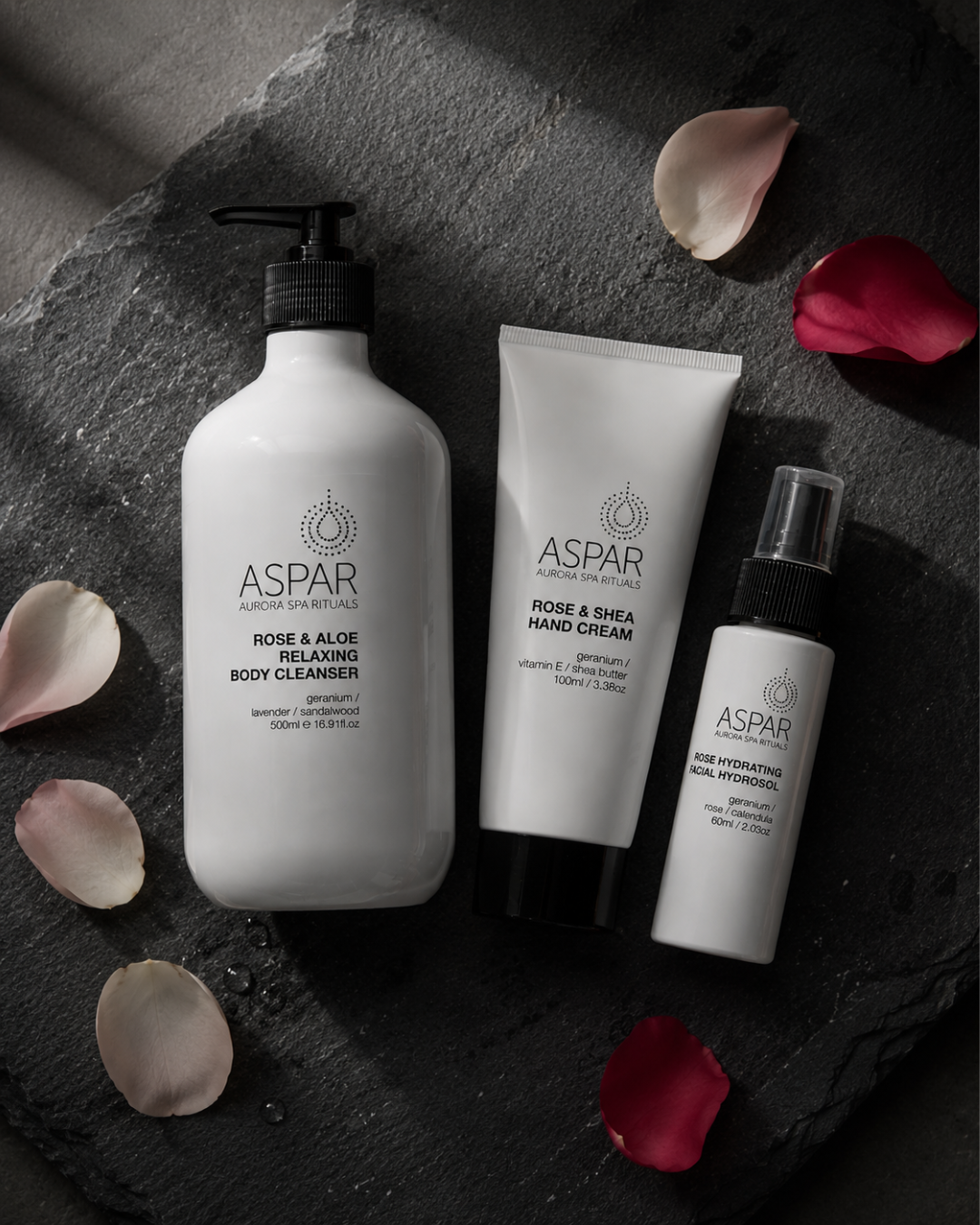 Three ASPAR Advanced Dermatology body care products arranged on a dark stone surface with scattered rose petals: a large white pump bottle of Rose & Aloe Relaxing Body Cleanser, a white squeeze tube of Rose & Shea Hand Cream, and a smaller white spray bottle of Rose Hydrating Hand Cream.