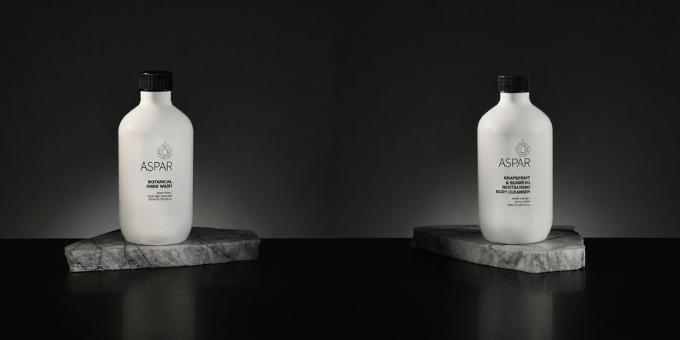 Two white ASPAR shampoo bottles with black caps, displayed side-by-side on rectangular gray stone slabs against a dark black background.