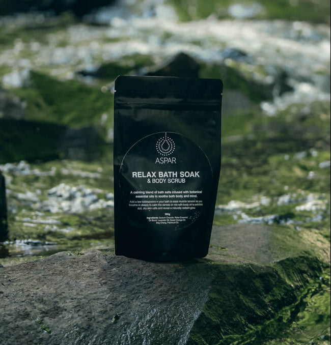 Black packaging of 'Relax Bath Soak & Body Scrub' by ASPAR on a natural stone background.
