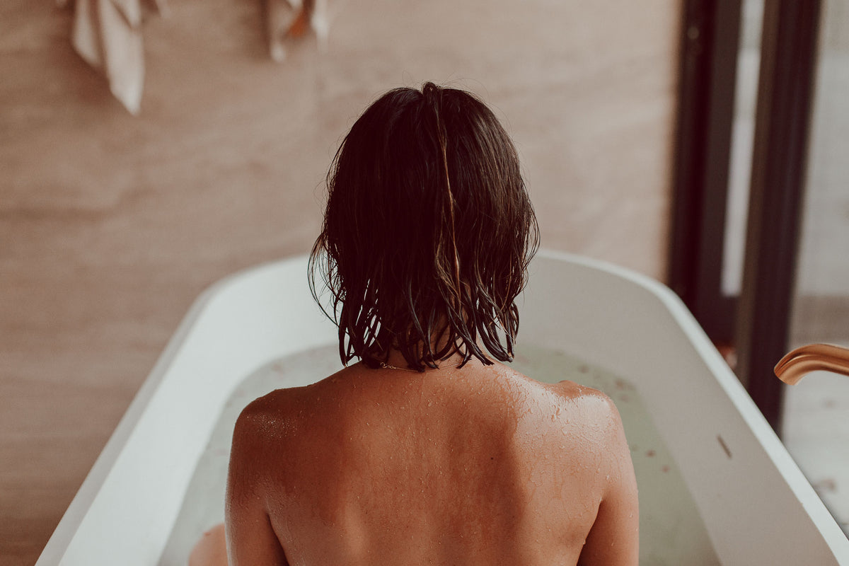 A person with wet dark hair sits in a white bathtub viewed from behind, with a warm-toned bathroom background and wooden door frame visible.