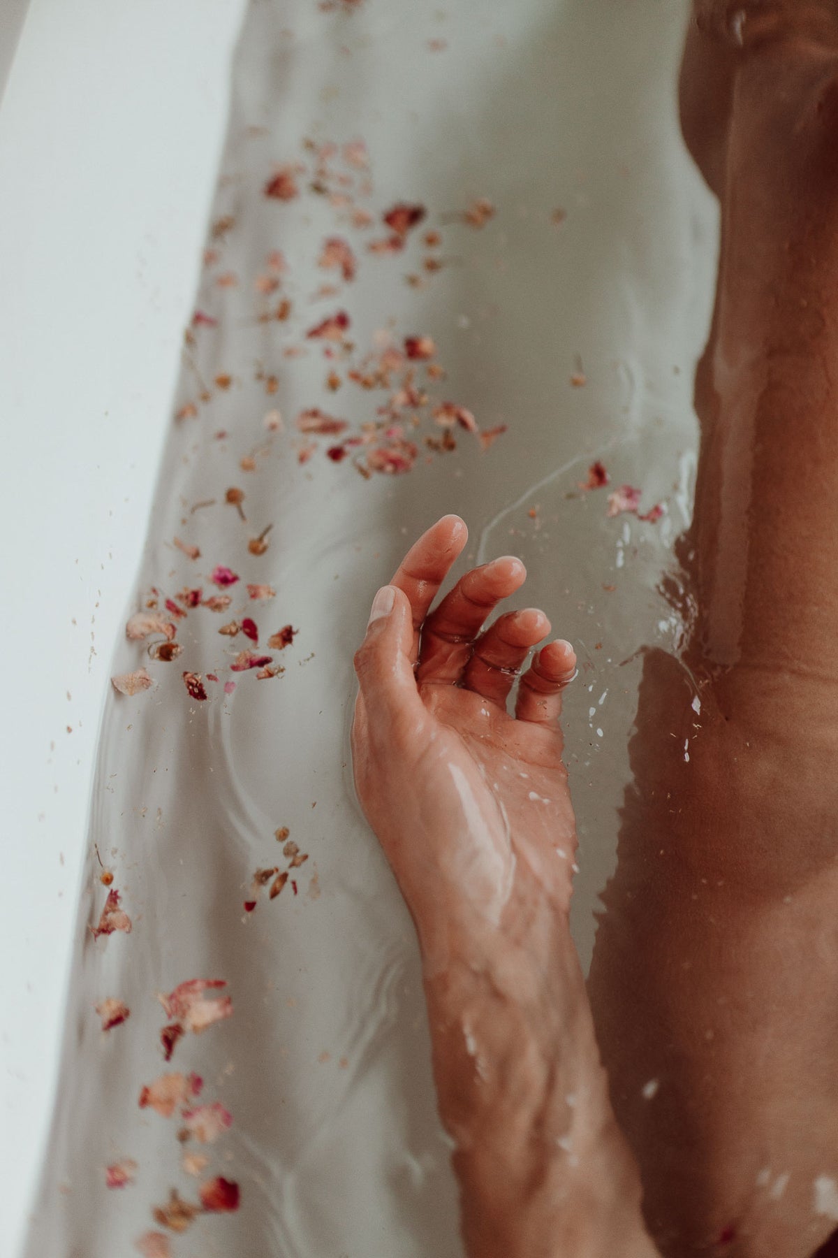A close-up lifestyle shot of a person's hand resting in clear, bubbly bath water with scattered dried rose petals floating around.
