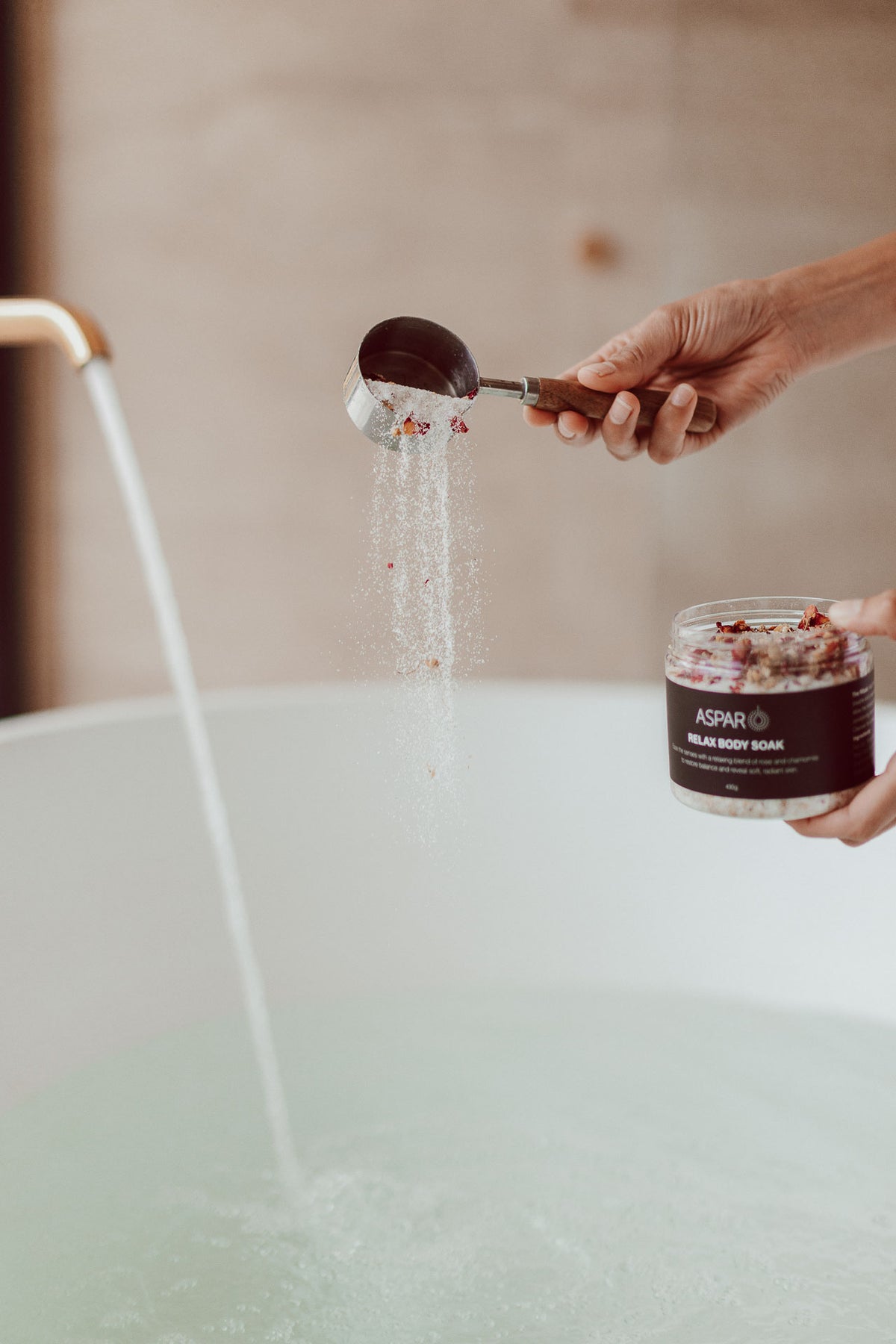 A person holds a jar of ASPAR Relax Body Soak and a metal scoop filled with the powder over a running faucet into a bathtub.