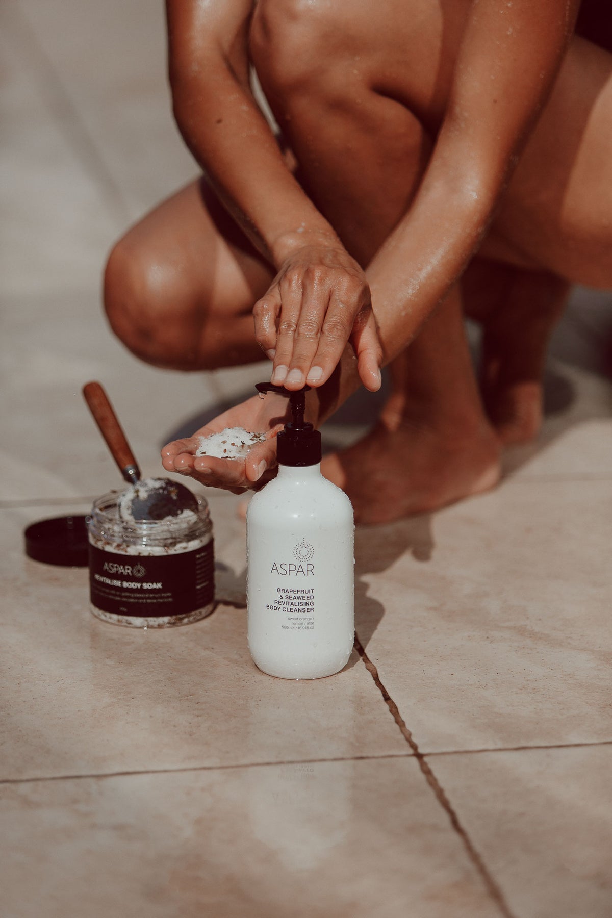 A person kneels on a tiled floor applying a white scrub from a small jar into their hand, with a larger white ASPAR Body Cleanser bottle standing nearby.