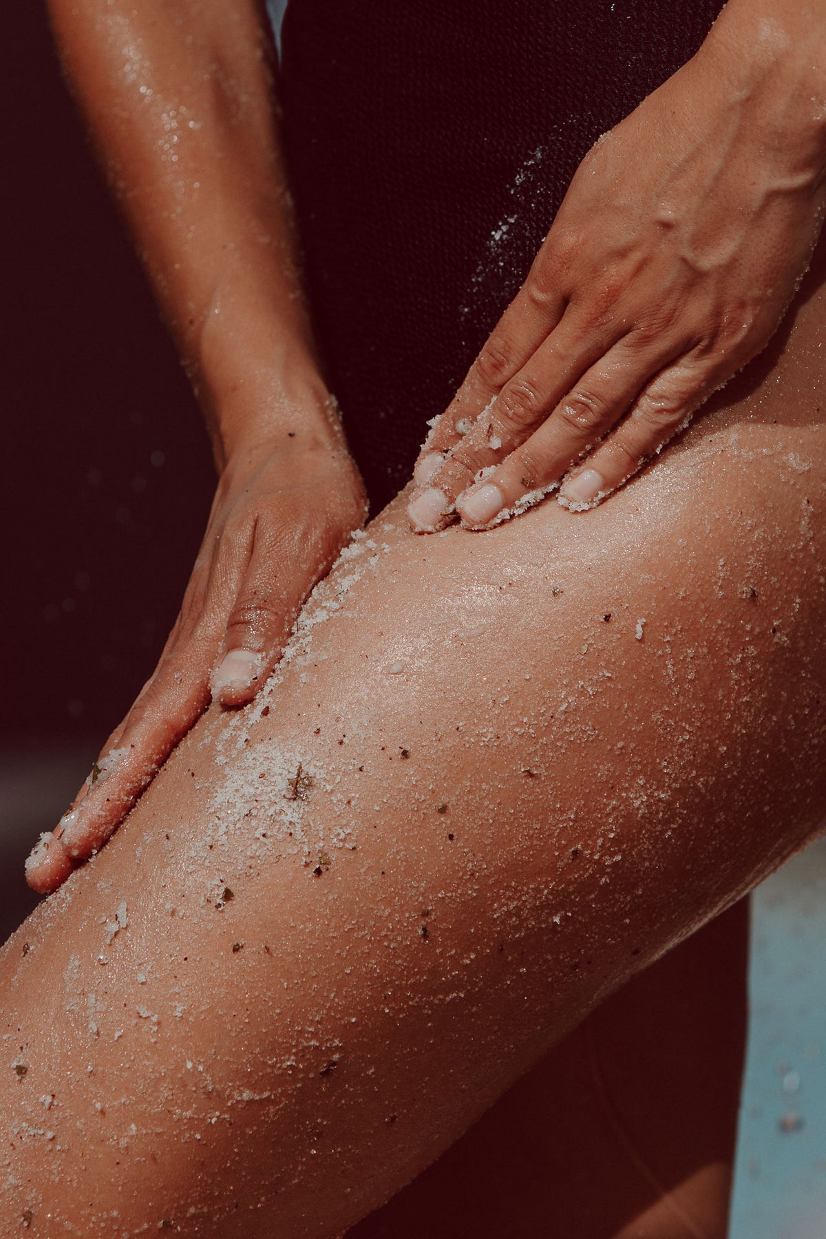 Close-up of a person's leg and hands applying or rubbing a white textured scrub or sand-like product onto the skin.