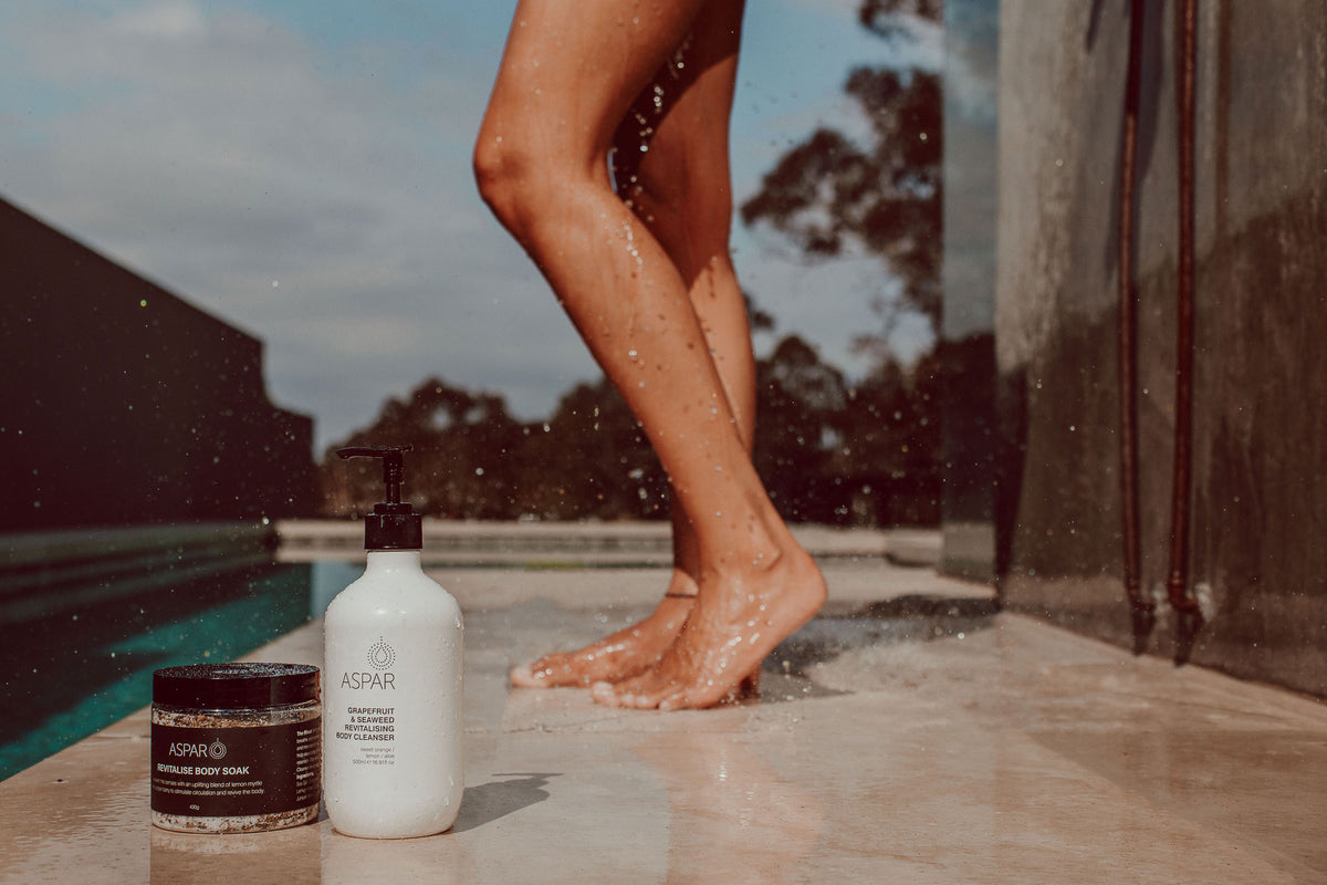 A lifestyle shot of ASPAR body care products on a poolside deck with water droplets splashing. In the foreground, a white bottle of Grapefruit & Seaweed Body Cleanser stands next to a dark lid clear jar of Revitalise Body Soak.