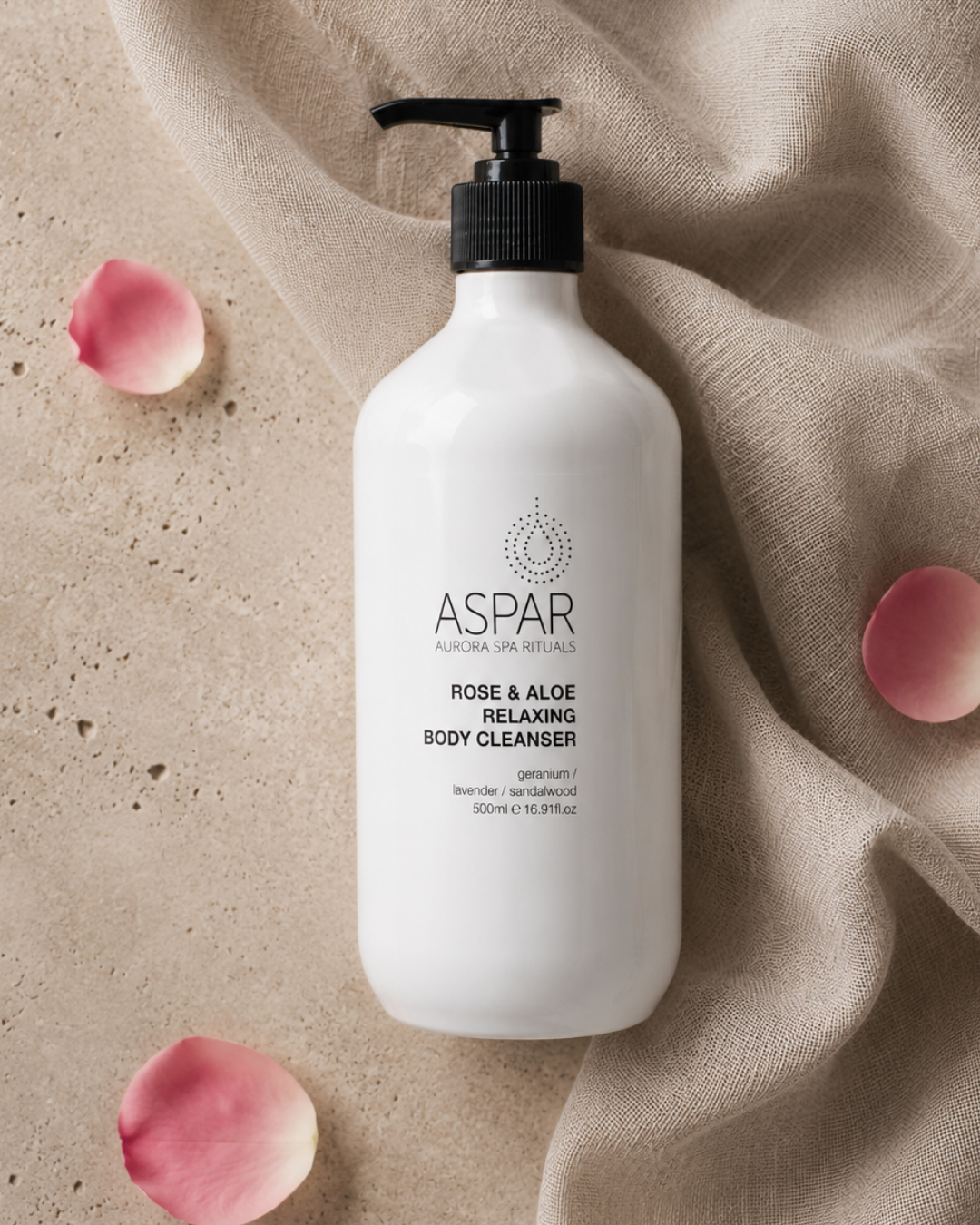 A 500ml white pump bottle of ASPAR Rose & Aloe Relaxing Body Cleanser, styled on a textured beige stone surface with draped linen fabric and pink rose petals.