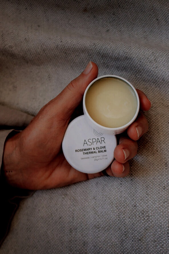 Hand holding a container of Aspar balm on a textured fabric background