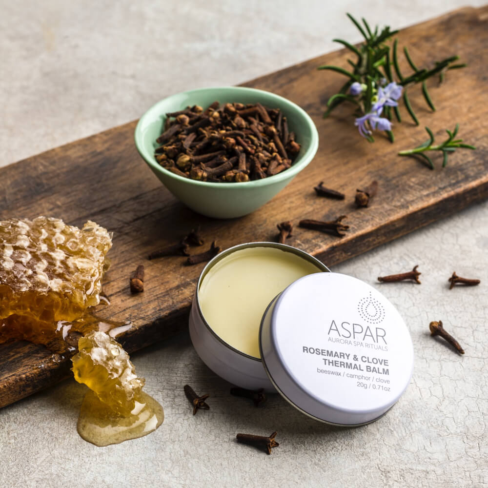 Aspar rosemary and clove thermal balm on a wooden board with honeycomb and cloves.