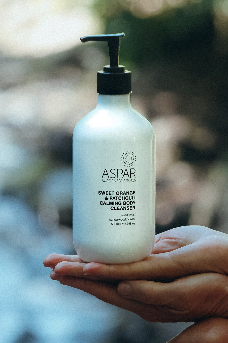 Hand holding a bottle of Aspar Sweet Orange & Patchouli Calming Body Cleanser with a blurred natural background.