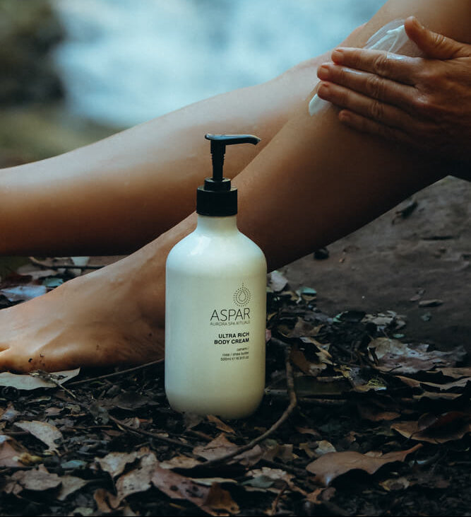 Person applying body cream to their leg with a bottle of Aspar cream on a natural setting.