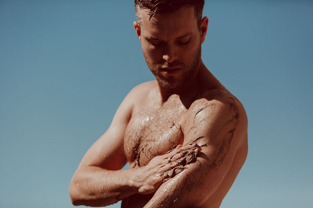A shirtless man outdoors with ASPAR body scrub applied to his body, looking downward against a clear blue sky.